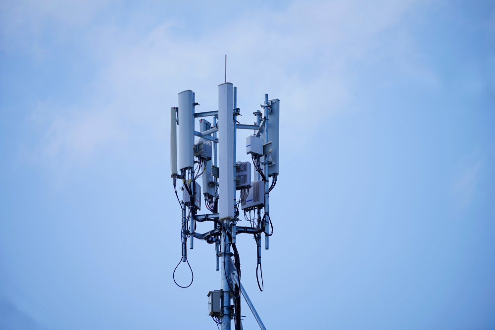 A cell tower with multiple antennas and transceiver units against a clear blue sky, likely utilizing frequency stabilization technology like TCXOs for communication signals.